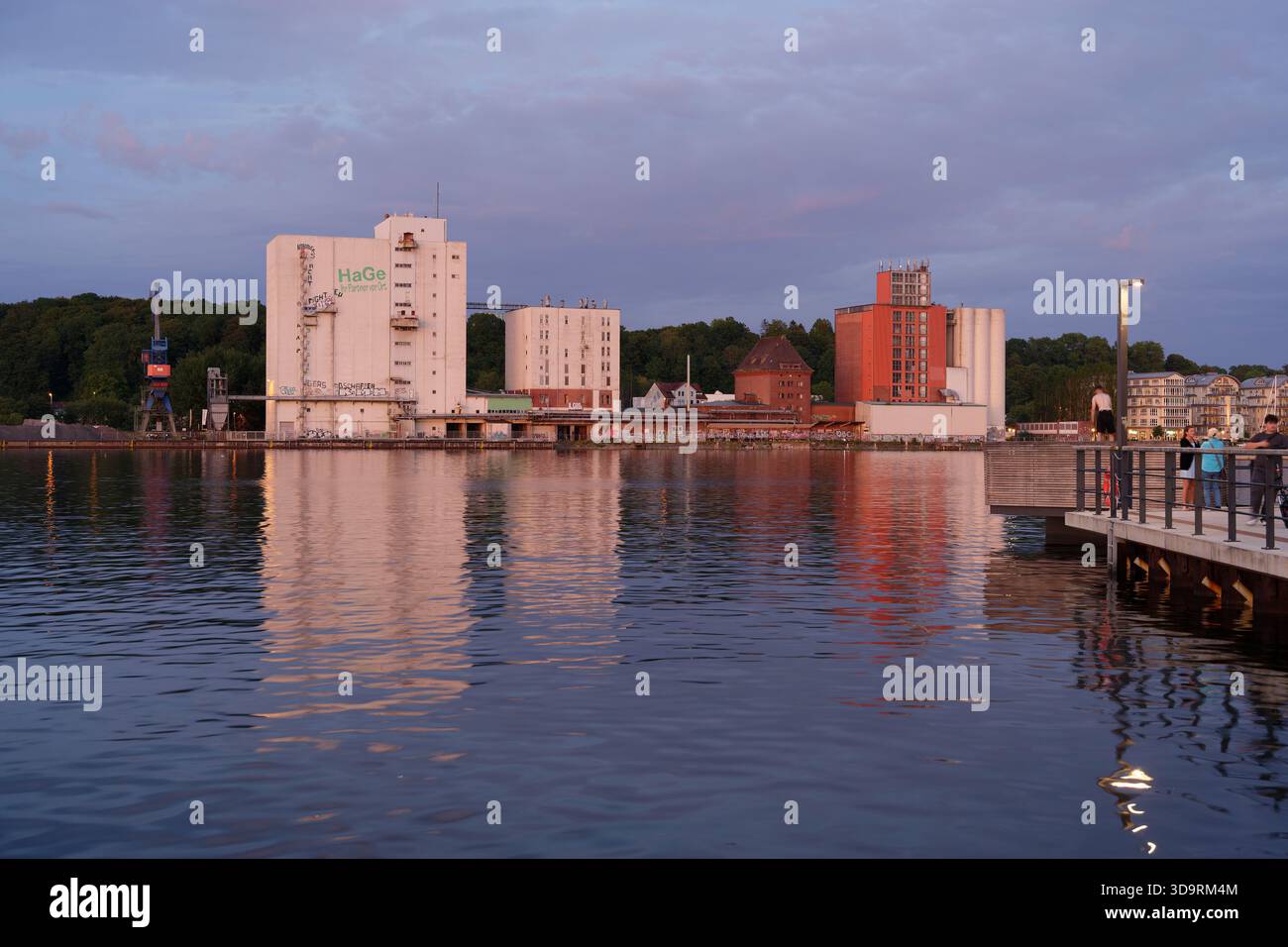 Un'affascinante città portuale presenta una passeggiata sul lungomare lungo l'acqua. In lontananza, sorge un edificio industriale che mescola l'attività urbana di Flensb Foto Stock