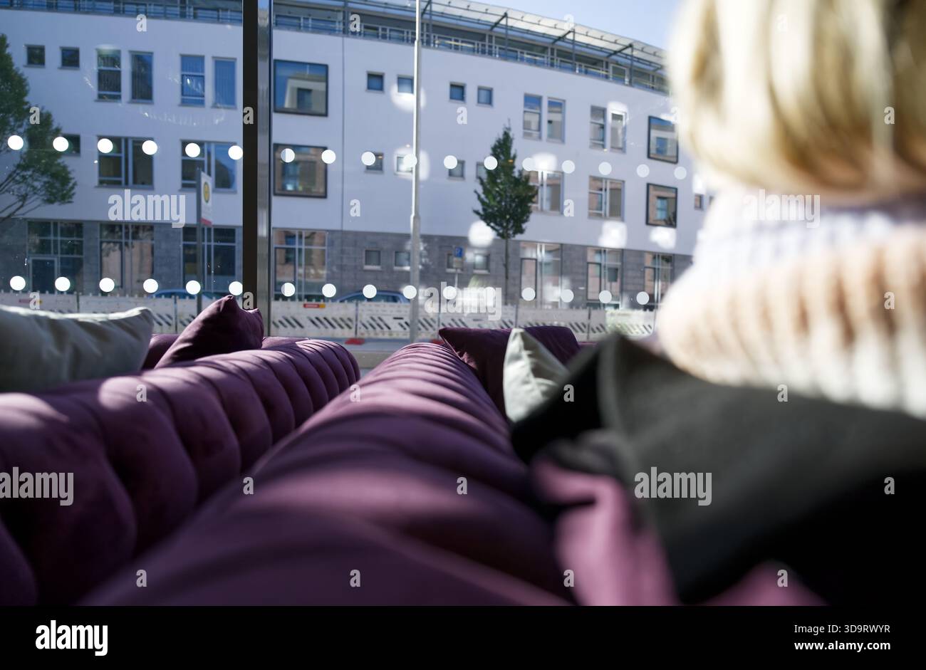 Un ambiente interno accogliente con un morbido divano viola e una vista di un edificio moderno attraverso grandi finestre. Una donna bionda siede sul divano; l'ambiente è b Foto Stock