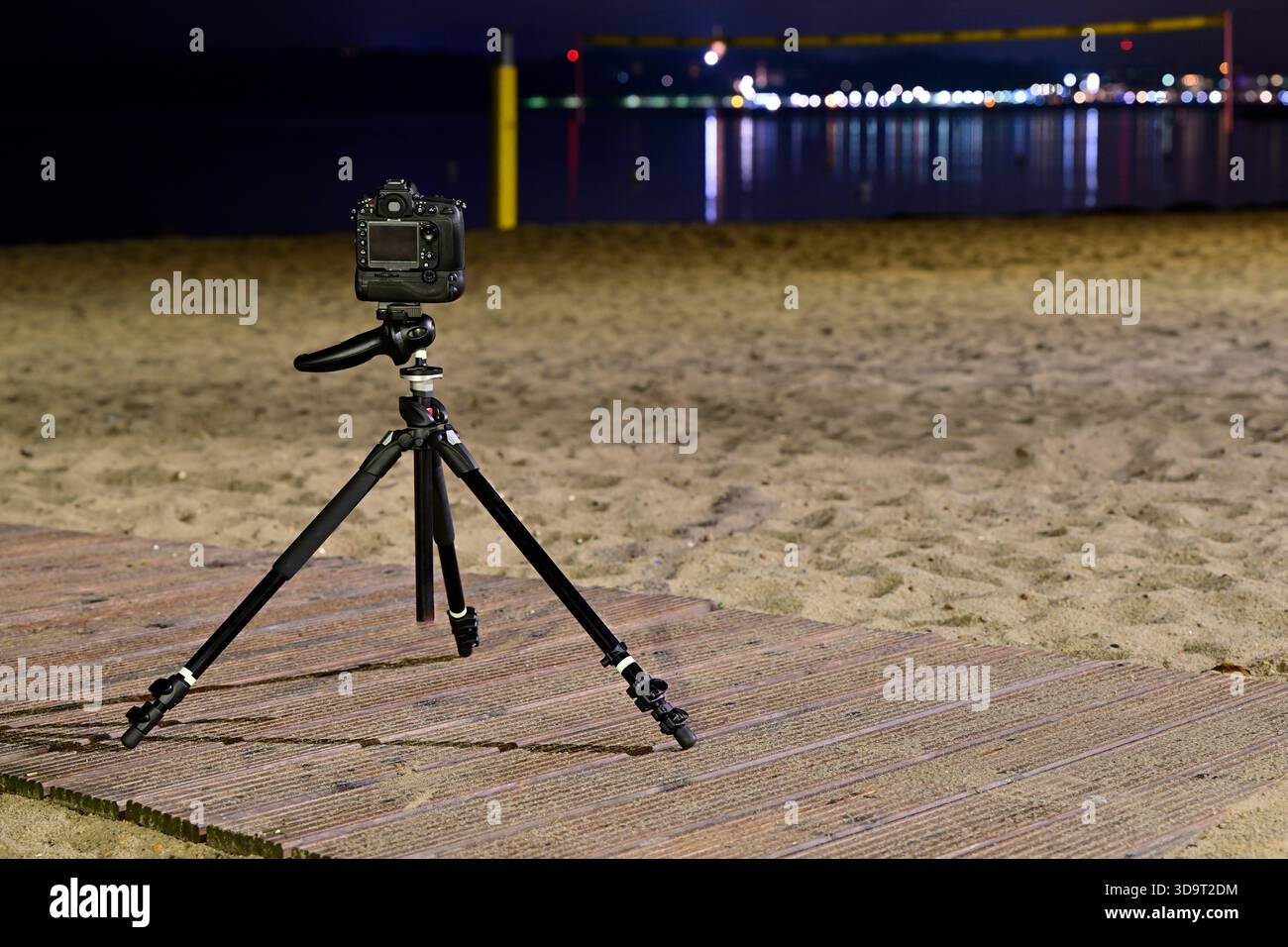Una fotocamera montata su un treppiede posizionato su una spiaggia sabbiosa di notte, con una superficie d'acqua calmo che riflette luci lontane e una rete da pallavolo nel bac Foto Stock