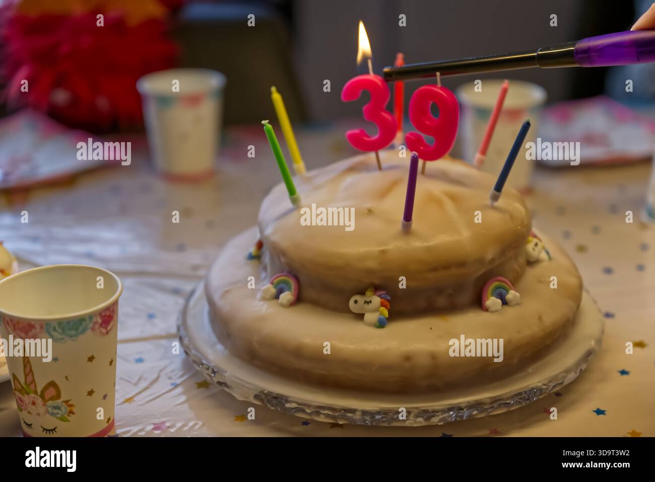 Una torta di compleanno con il numero 39 in cima, circondata da candele colorate. La torta è decorata con piccole figure arcobaleno e unicorno, set o Foto Stock
