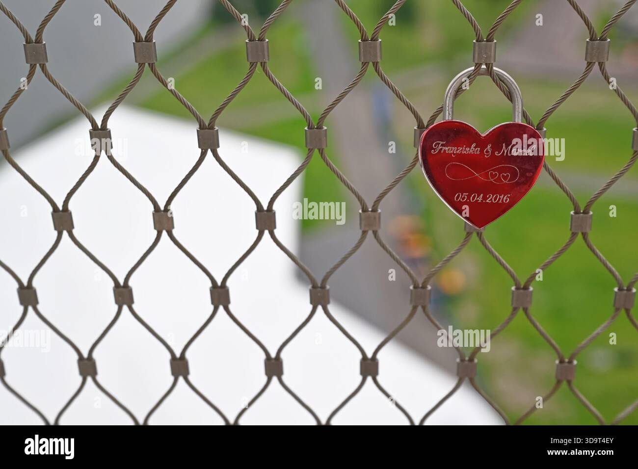 Un lucchetto rosso a forma di cuore è appeso a una recinzione in rete metallica, con incisi nomi e una data. Lo sfondo presenta un paesaggio verde sfocato, che suggerisce Foto Stock