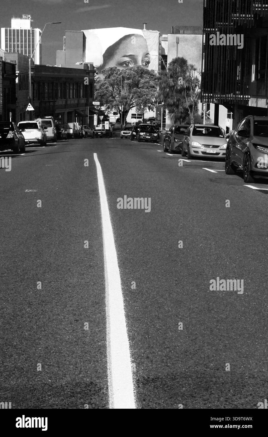 Edificio murale di un volto, sotto gli occhi oscurati da un albero, guardando lungo una linea bianca della strada verso gli occhi alla fine della strada Foto Stock