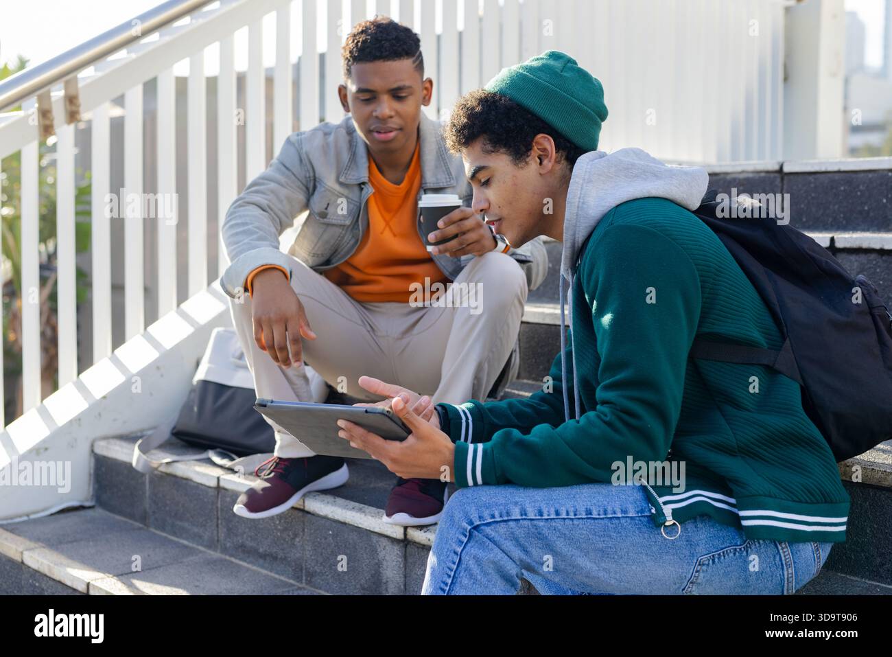 Amici afroamericani seduti ai gradini del campus usando un tablet, tenendo una tazza di caffè accanto agli zaini Foto Stock