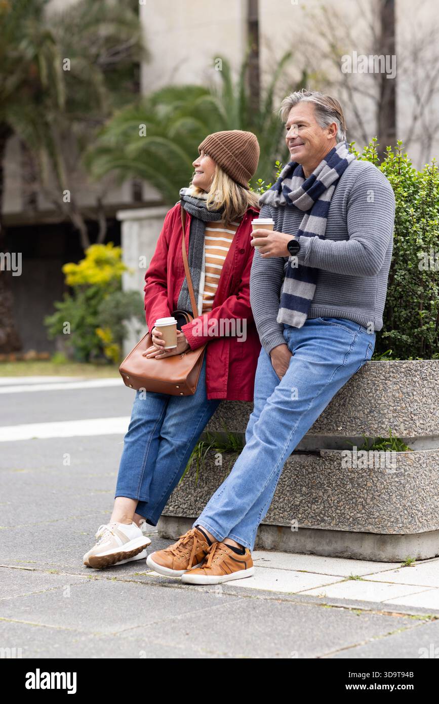 La coppia senior seduto sulla panchina della piantatrice in cemento in piazza regge tazze da caffè e indossa uno smartwatch Foto Stock