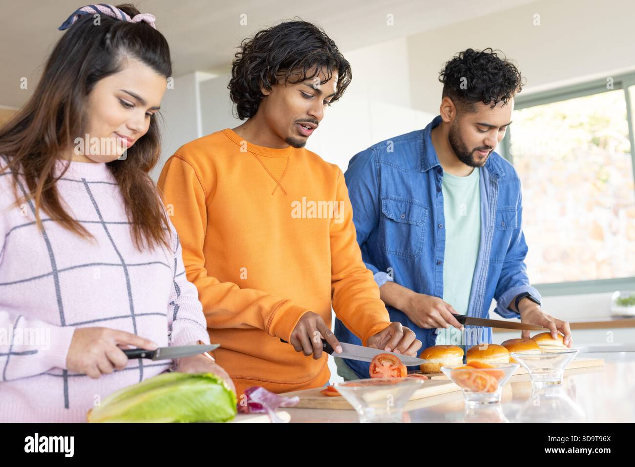 Diversi amici affettano i panini di pomodoro con lattuga in ciotole di vetro sui taglieri al banco della cucina Foto Stock