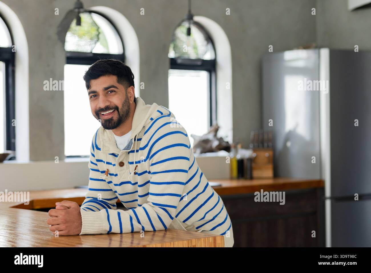 Uomo indiano appoggiato su un piano di cucina in legno, sorridente accanto alle bottiglie di olio d'oliva e al frigorifero Foto Stock