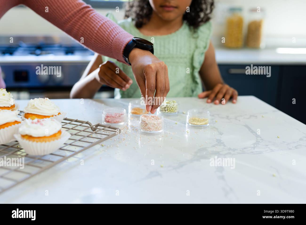 Mano di un adulto che tiene il vaso sprinkles sulla parte superiore in marmo, mano del bambino che riposa su una griglia con cupcake smerigliati Foto Stock