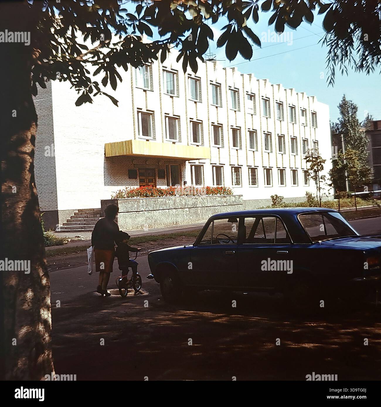 Fotografia d'archivio vintage degli anni '1980 che cattura l'esterno della scuola di musica per bambini a Sloviansk, URSS. L'edificio in mattoni bianchi presenta una distintiva facciata ritmica con pilastri verticali e un baldacchino d'ingresso oro. Una berlina sovietica blu è parcheggiata per strada in primo piano. Incorniciata da rami d'albero, la scena mostra una donna con un bambino vicino all'ingresso. Questa immagine autentica riflette l'attenzione sull'educazione culturale e la tranquilla atmosfera quotidiana nella regione del Donbas durante la fine dell'era sovietica. Foto Stock