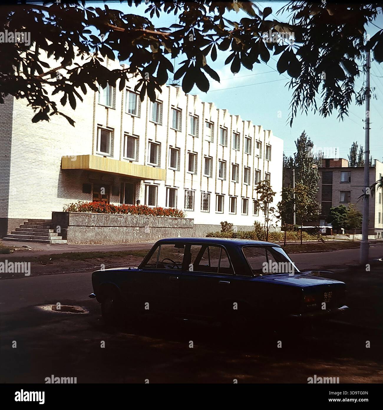 Fotografia d'archivio vintage degli anni '1980 che cattura l'esterno della scuola di musica per bambini a Sloviansk, URSS. L'edificio in mattoni bianchi presenta una distintiva facciata ritmica con pilastri verticali e un baldacchino d'ingresso oro. Una berlina sovietica blu è parcheggiata per strada in primo piano. Incorniciata da rami d'albero, la scena mostra una donna con un bambino vicino all'ingresso. Questa immagine autentica riflette l'attenzione sull'educazione culturale e la tranquilla atmosfera quotidiana nella regione del Donbas durante la fine dell'era sovietica. Foto Stock