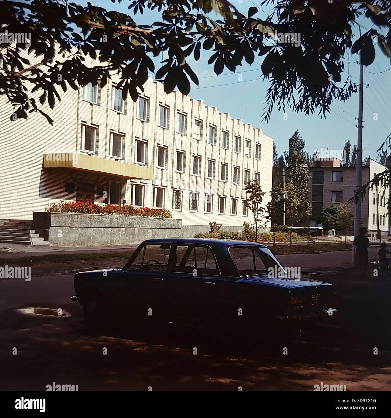 Fotografia d'archivio vintage degli anni '1980 che cattura l'esterno della scuola di musica per bambini a Sloviansk, URSS. L'edificio in mattoni bianchi presenta una distintiva facciata ritmica con pilastri verticali e un baldacchino d'ingresso oro. Una berlina sovietica blu è parcheggiata per strada in primo piano. Incorniciata da rami d'albero, la scena mostra una donna con un bambino vicino all'ingresso. Questa immagine autentica riflette l'attenzione sull'educazione culturale e la tranquilla atmosfera quotidiana nella regione del Donbas durante la fine dell'era sovietica. Foto Stock