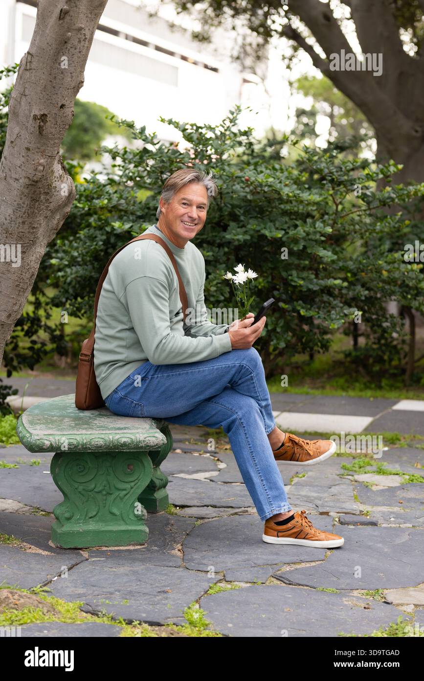 Uomo di mezza età seduto su una panchina di pietra verde nel parco urbano con smartphone e fiori bianchi Foto Stock