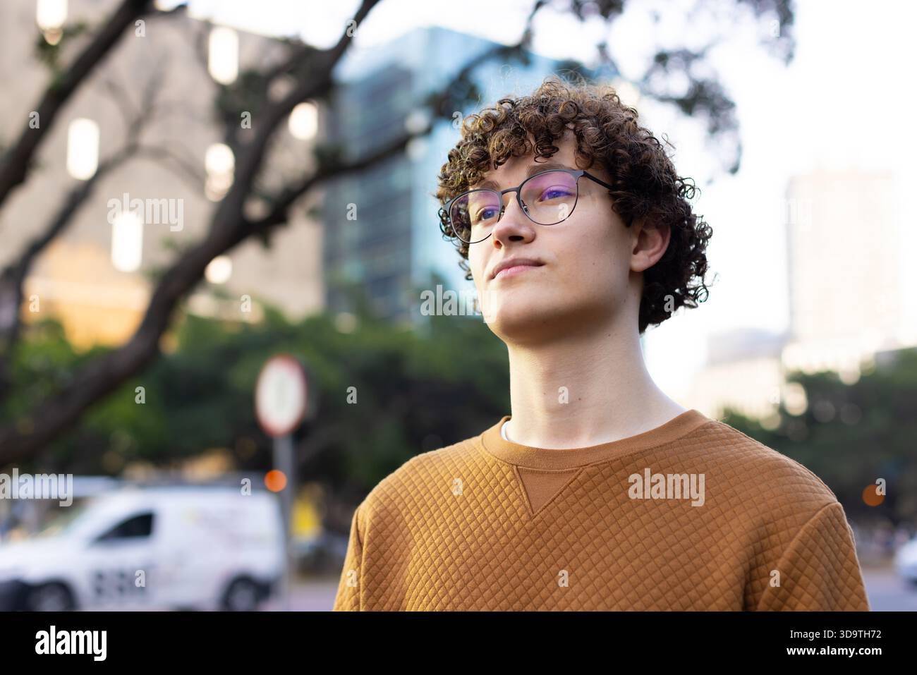 Studente maschio che indossa una felpa trapuntata e occhiali da vista rotondi mentre guarda verso l'alto sul marciapiede della città Foto Stock