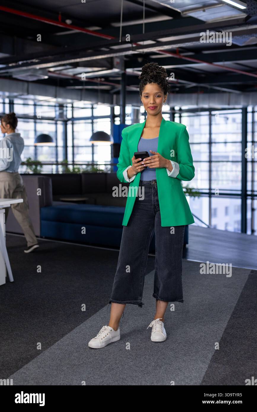 Colleghi diversi che passano accanto alla sala d'ufficio mentre uno indossa un blazer verde e utilizza lo smartphone Foto Stock