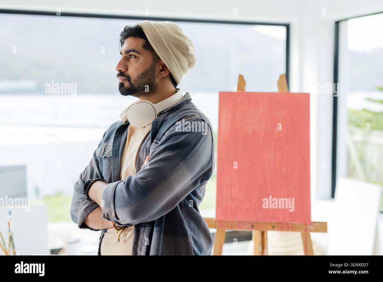 Uomo indiano in piedi accanto alla finestra dello studio con le cuffie, che guarda il lago con tela rossa e cavalletto Foto Stock