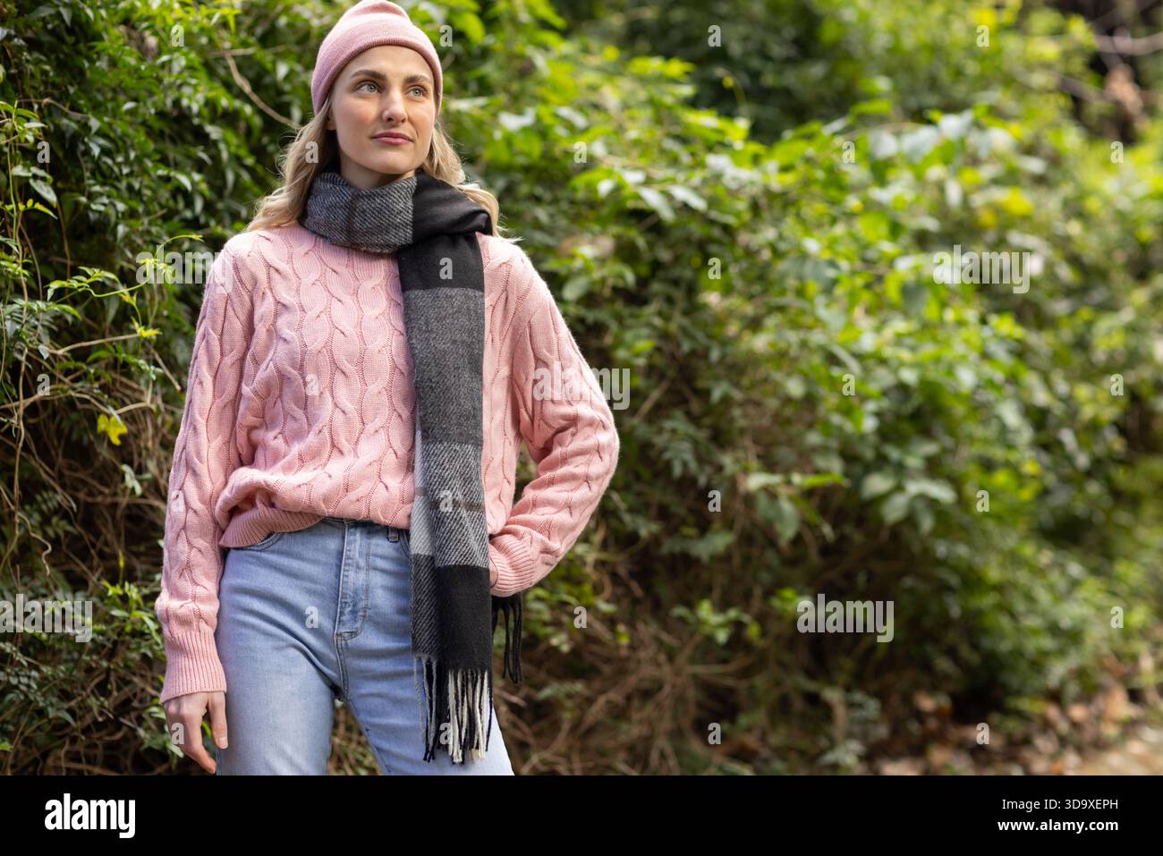 Donna in piedi nel parco accanto alla siepe che indossa un maglione in maglia rosa, sciarpa a righe e berretto, spazio per copiare Foto Stock