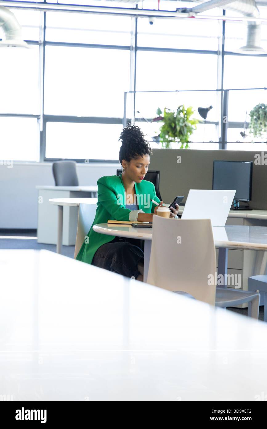 Donna afroamericana che controlla smartphone e jotting notes nel notebook di un ufficio open space Foto Stock