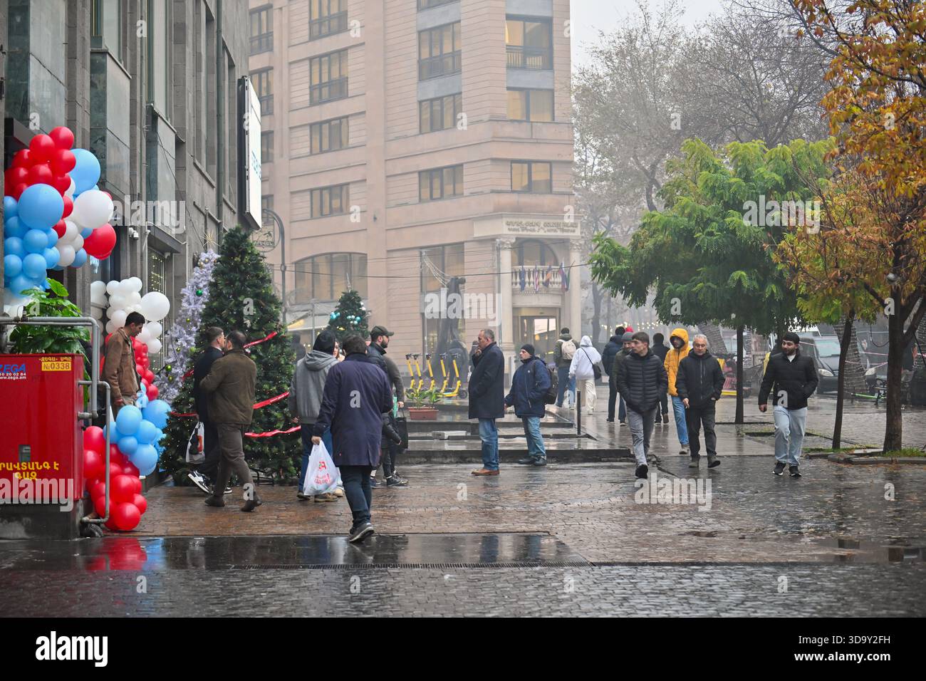 Erevan Armenia 12.7,2025. Un gruppo di persone sta camminando lungo una strada bagnata di fronte a un edificio. La strada è coperta di pozzanghere e la gente lo è Foto Stock
