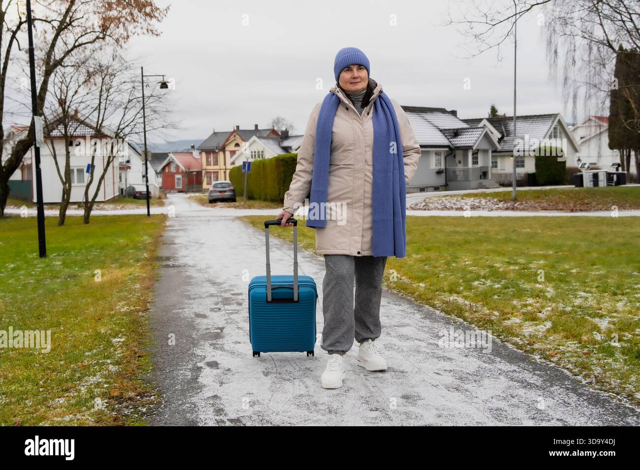Viaggiatrice di mezza età con valigia blu si erge su un sentiero innevato, vestito con caldo cappotto e sciarpa, con case residenziali sullo sfondo, capturi Foto Stock