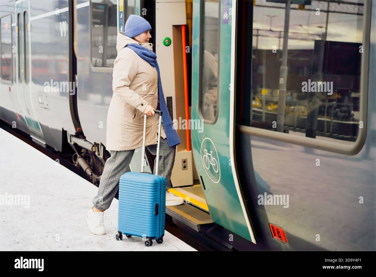 La donna di mezza età che viaggia con la valigia blu si imbarcherà in treno alla stazione, vestita con un caldo abbigliamento invernale, catturando l'essenza del viaggio e dell'anticipo Foto Stock