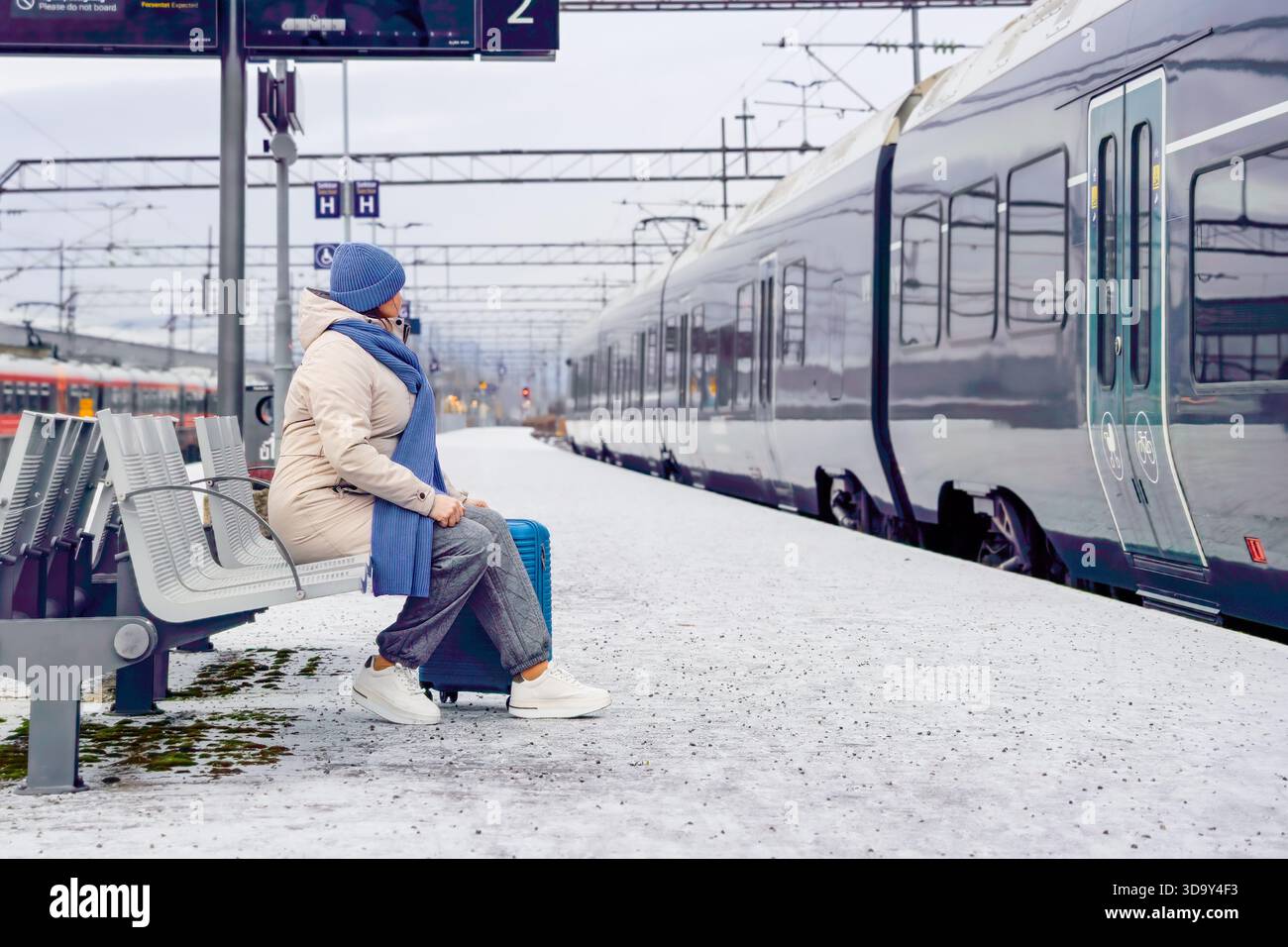 Donna matura viaggiatrice con valigia siede su una panchina in una stazione ferroviaria innevata, in attesa dell'arrivo del treno, vestita con caldi abiti invernali, catturando la bellezza Foto Stock