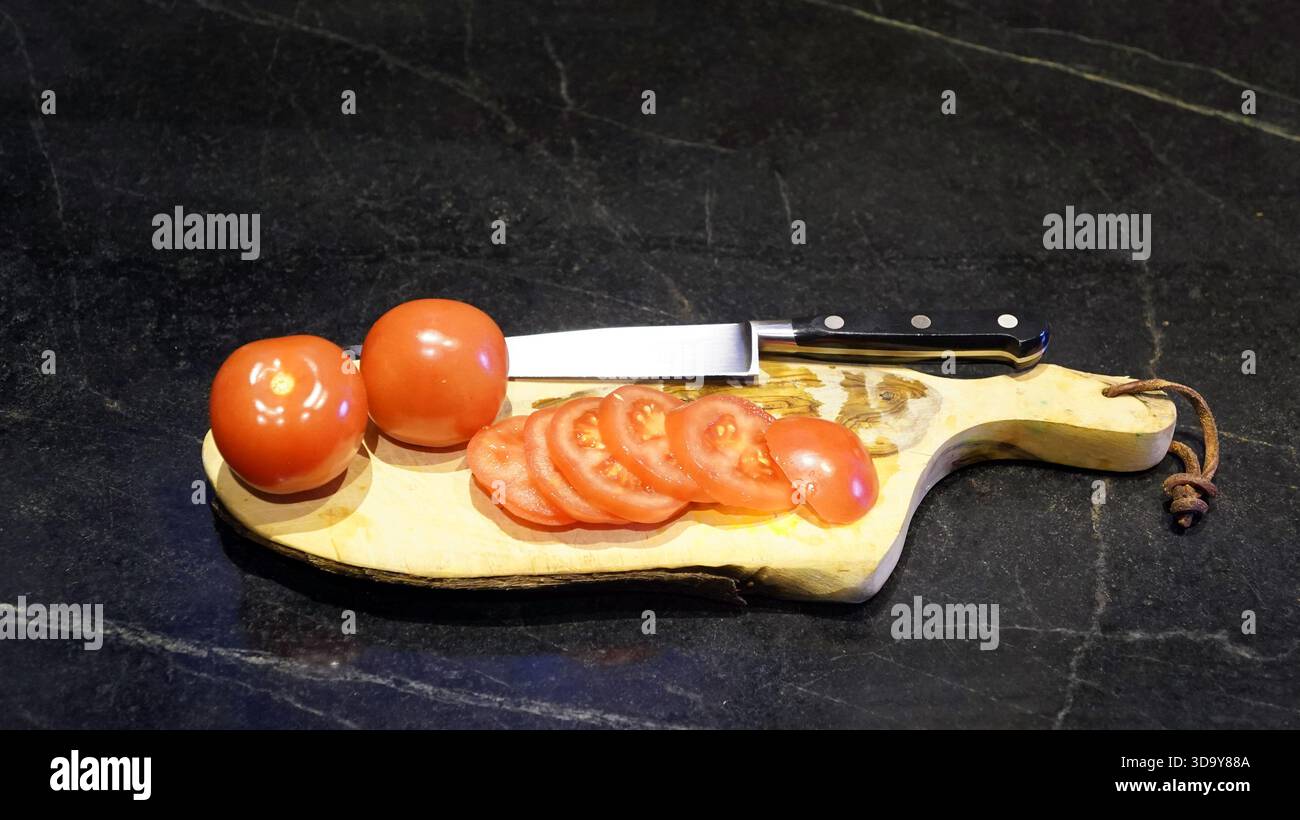Un piccolo tagliere di legno che contiene due pomodori rossi interi, una serie di fette di pomodoro fresco e un coltello da cucina argentato si trova in cima a un piano scuro Foto Stock