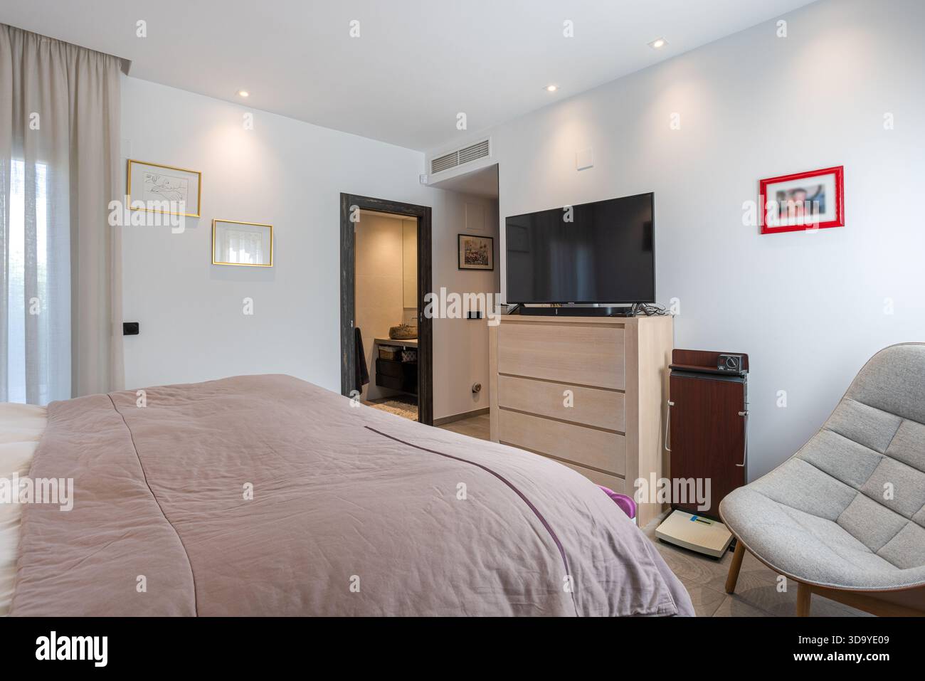 Accogliente camera da letto con televisione, comò, poltrona e soffice biancheria da letto in malva Foto Stock