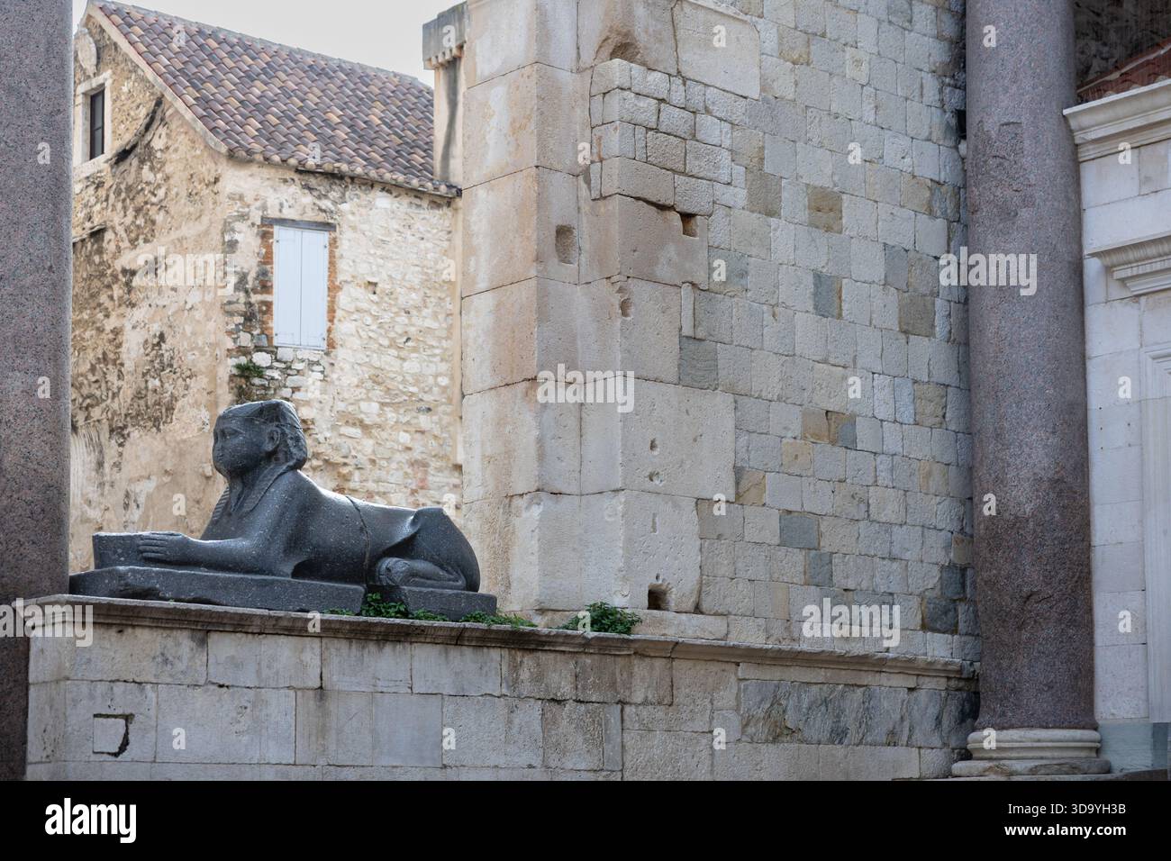 Un leone di pietra reclinabile intempestivo poggia su un piedistallo all'interno di un antico cortile. Pareti in pietra, colonne alte e un edificio rustico creano un'atmosfera senza tempo, Foto Stock