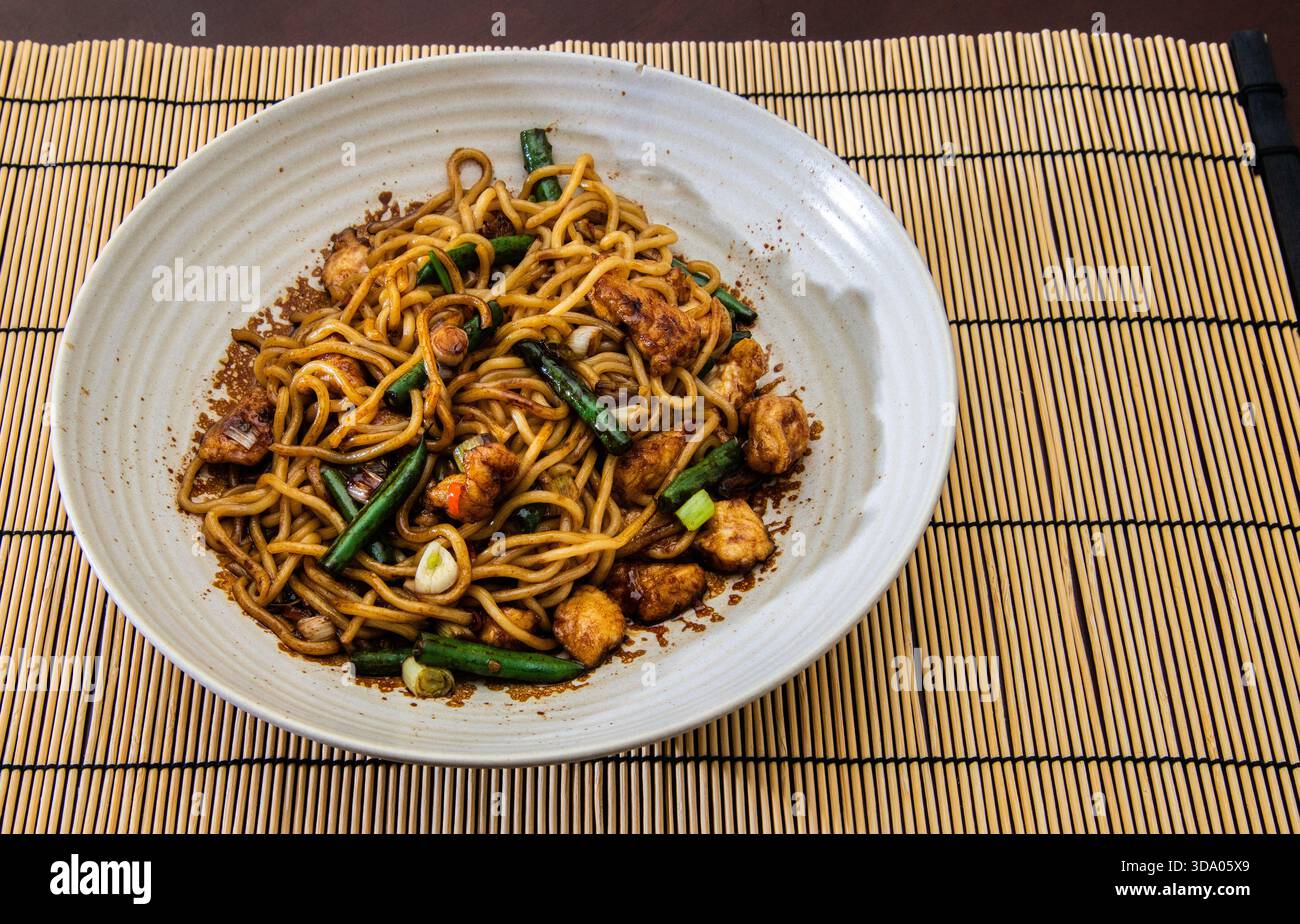 Una ciotola di noodles fritti al pollo su un tappetino di bambù, presentata in un ambiente pulito e sopraelevato. Foto Stock