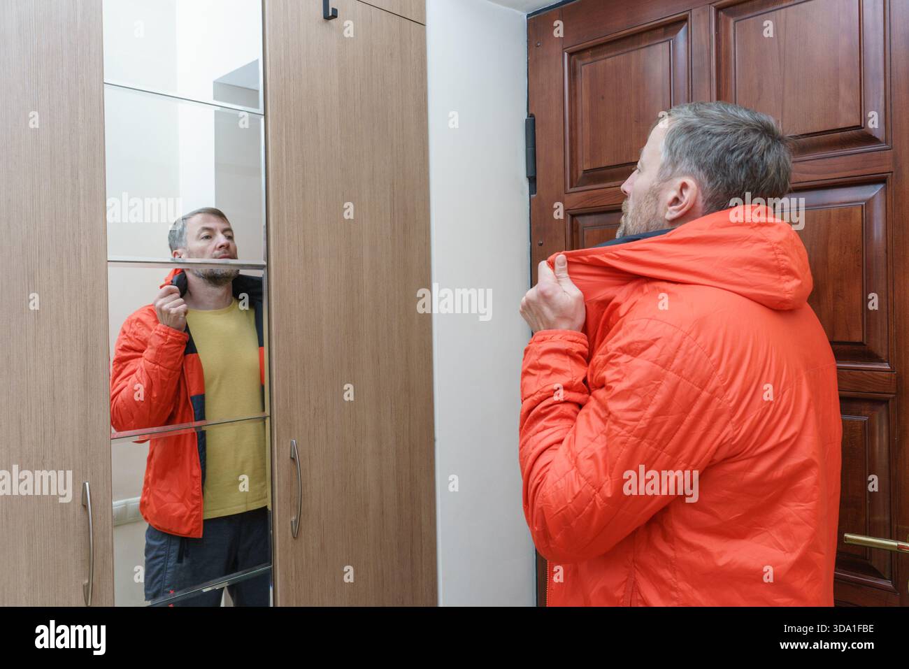 Un uomo di mezza età con la barba indossa una giacca arancione con cappuccio in un ingresso di casa, osservando il suo riflesso in una porta del guardaroba a specchio. Foto Stock