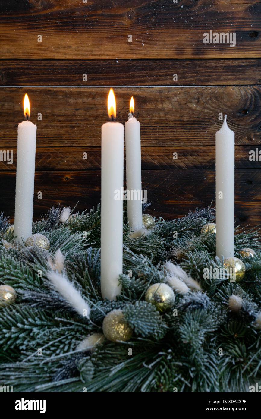 Tre alte candele bianche risplendono calorosamente sopra una corona sempreverde smerigliata decorata con ornamenti dorati. Foto Stock