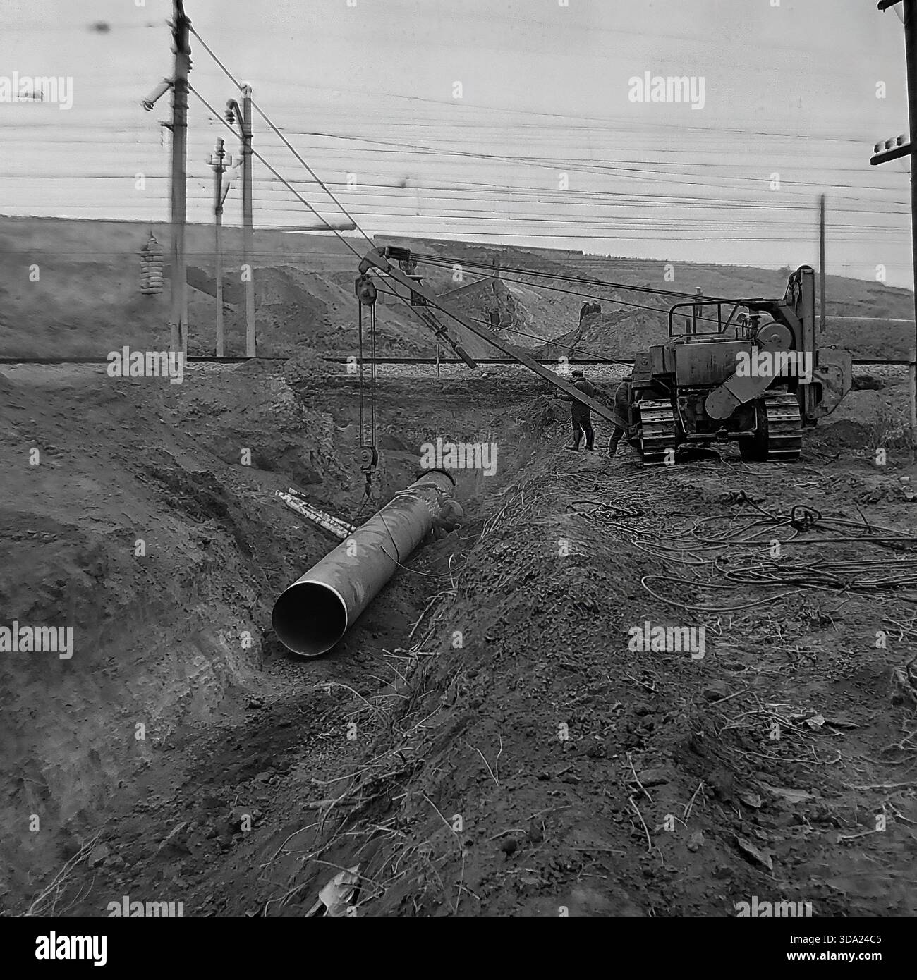 Regione di Donetsk, Ucraina, URSS. Foto dell'archivio storico degli anni '1970 della costruzione del gasdotto Shebelinka-Sloviansk vicino a una linea ferroviaria. Un posatubi cingolato sovietico pesante abbassa un grande tubo in acciaio in una profonda fossa accanto ai binari elettrificati. I lavoratori supervisionano l'installazione complessa in questa intersezione di infrastrutture critiche. Questa scena industriale evidenzia le enormi sfide ingegneristiche legate all'espansione della rete energetica nella steppa del Donbas durante l'era dell'economia pianificata. Foto Stock