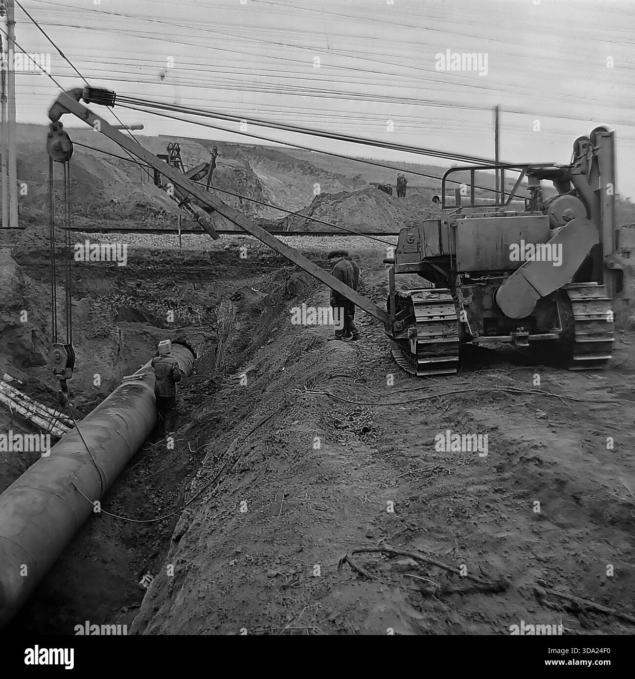 Regione di Donetsk, Ucraina, URSS. Foto dell'archivio storico degli anni '1970 della costruzione del gasdotto Shebelinka-Sloviansk vicino a una linea ferroviaria. Un posatubi cingolato sovietico pesante abbassa un grande tubo in acciaio in una profonda fossa accanto ai binari elettrificati. I lavoratori supervisionano l'installazione complessa in questa intersezione di infrastrutture critiche. Questa scena industriale evidenzia le enormi sfide ingegneristiche legate all'espansione della rete energetica nella steppa del Donbas durante l'era dell'economia pianificata. Foto Stock
