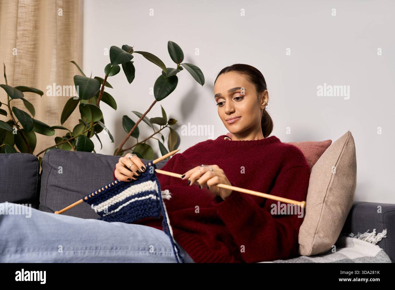 Una giovane donna afro-americana si diverte a lavorare a maglia in un accogliente maglione invernale a casa. Foto Stock