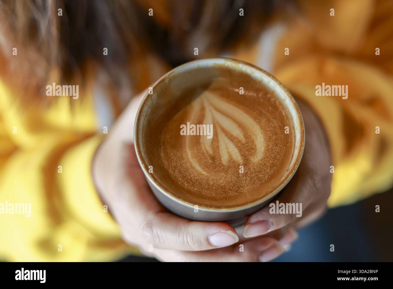 Donna che tiene a mano una tazza calda di caffè con latte art a forma di cuore. Vista dall'alto della donna che si gode momenti intimi con una bevanda calda, si sente rilassata e confortevole Foto Stock