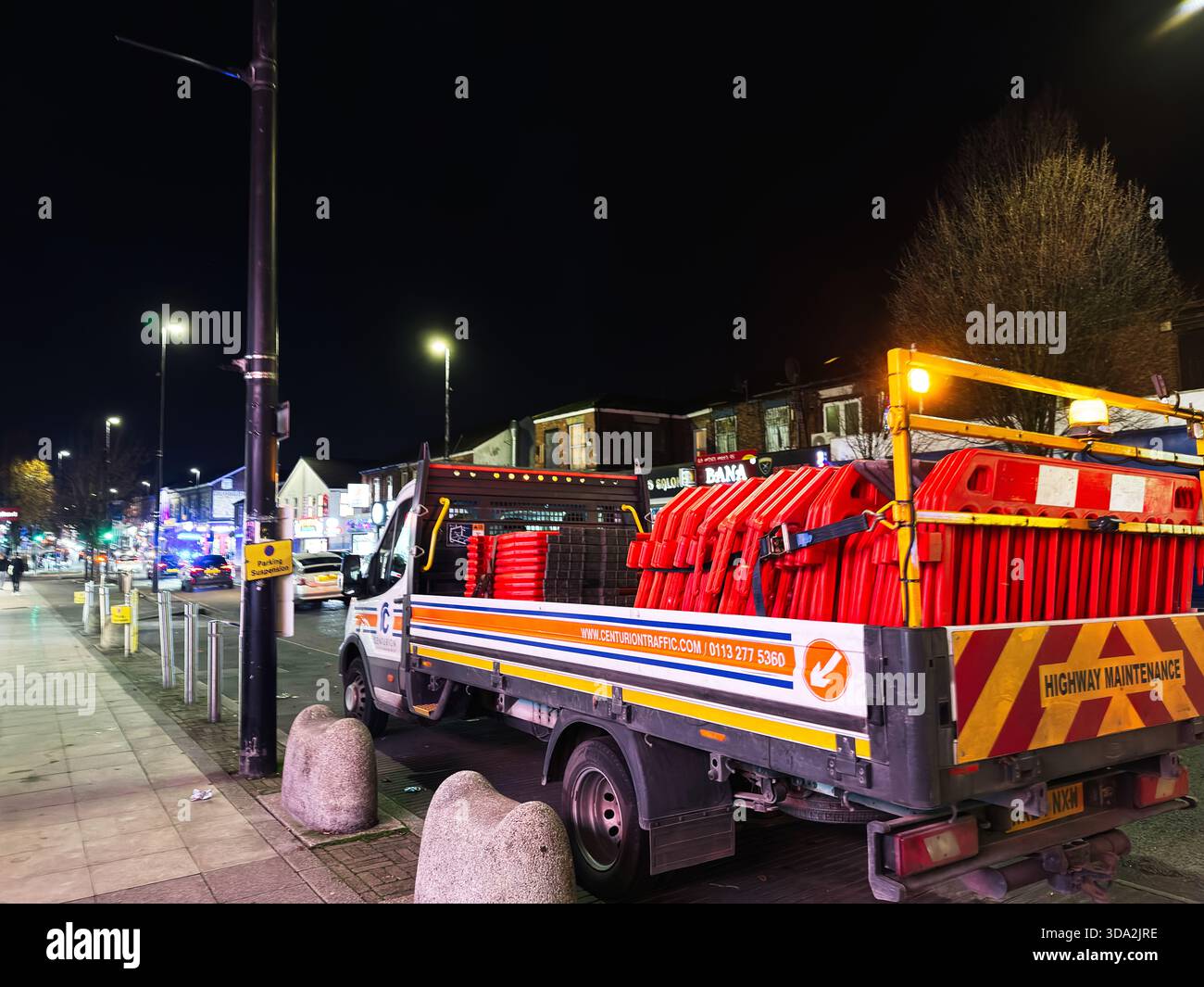 Salford, Regno Unito - 8 novembre 2025: La strada di Salford, notturna, mostra un camion per la manutenzione dell'autostrada carico di barriere rosse e luci luminose della città. Foto Stock