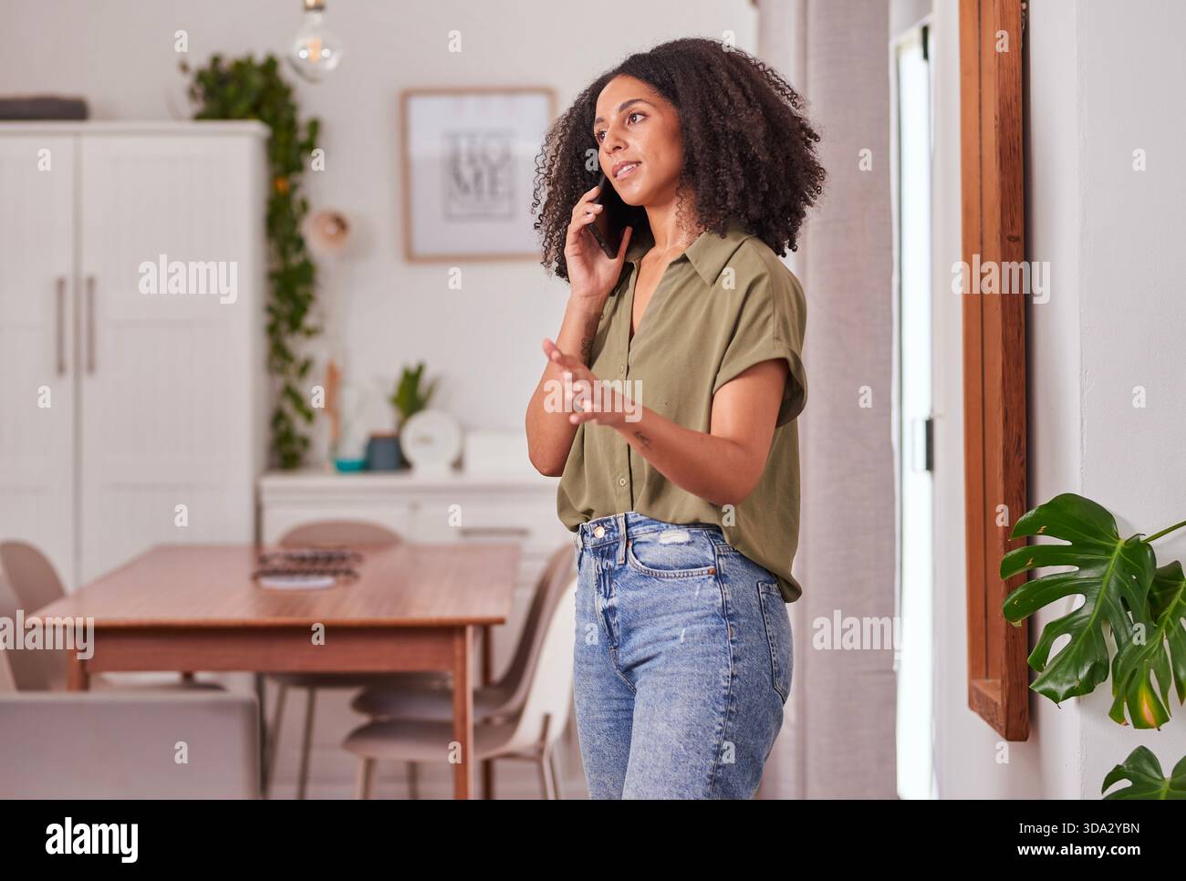 Donna informale al telefono negli interni moderni con piante e zona pranzo Foto Stock