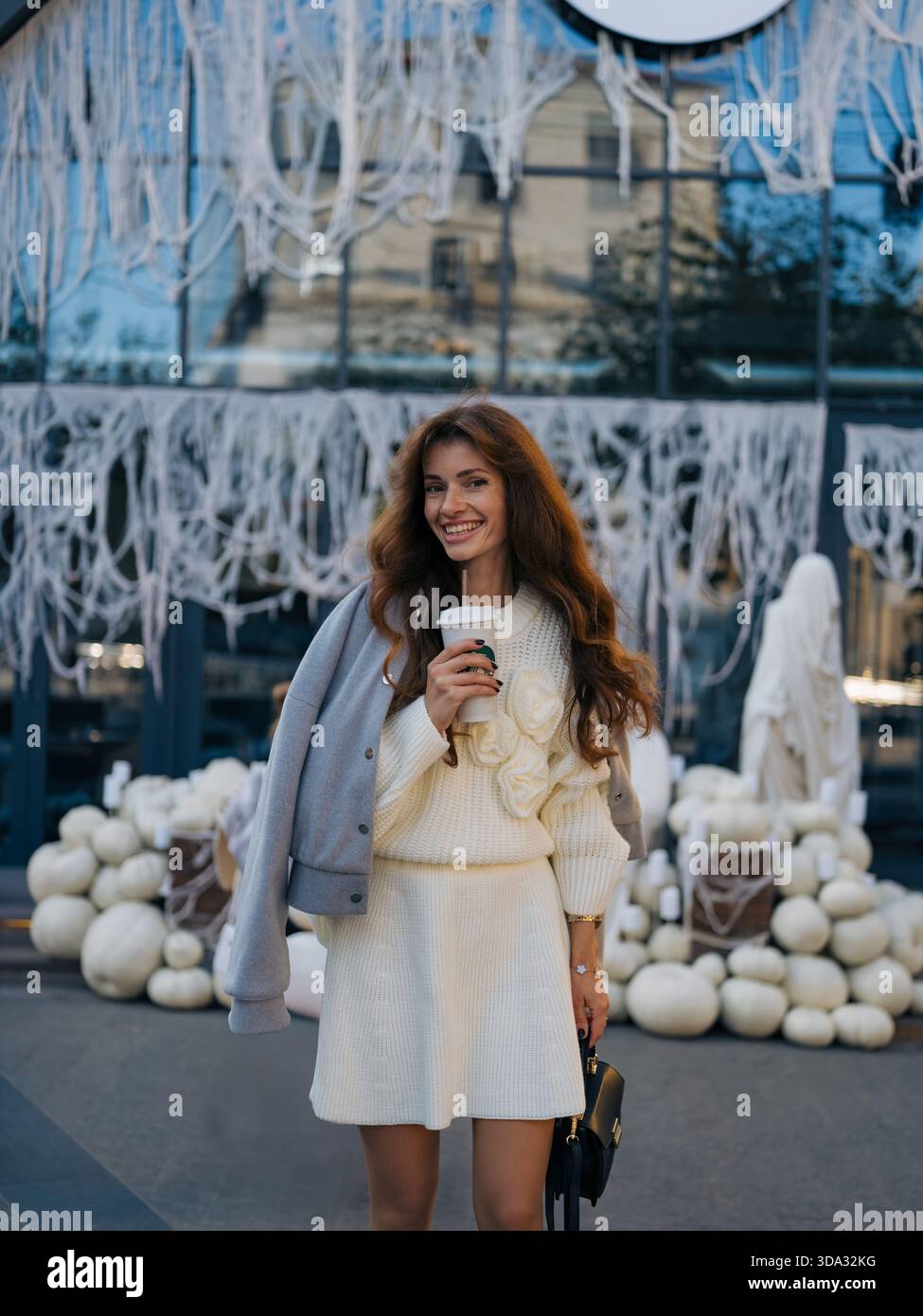 Donna alla moda in maglione bianco e giacca grigia in piedi all'aperto, sorridendo allegramente e tenendo il caffè contro il moderno Halloween bianco Foto Stock