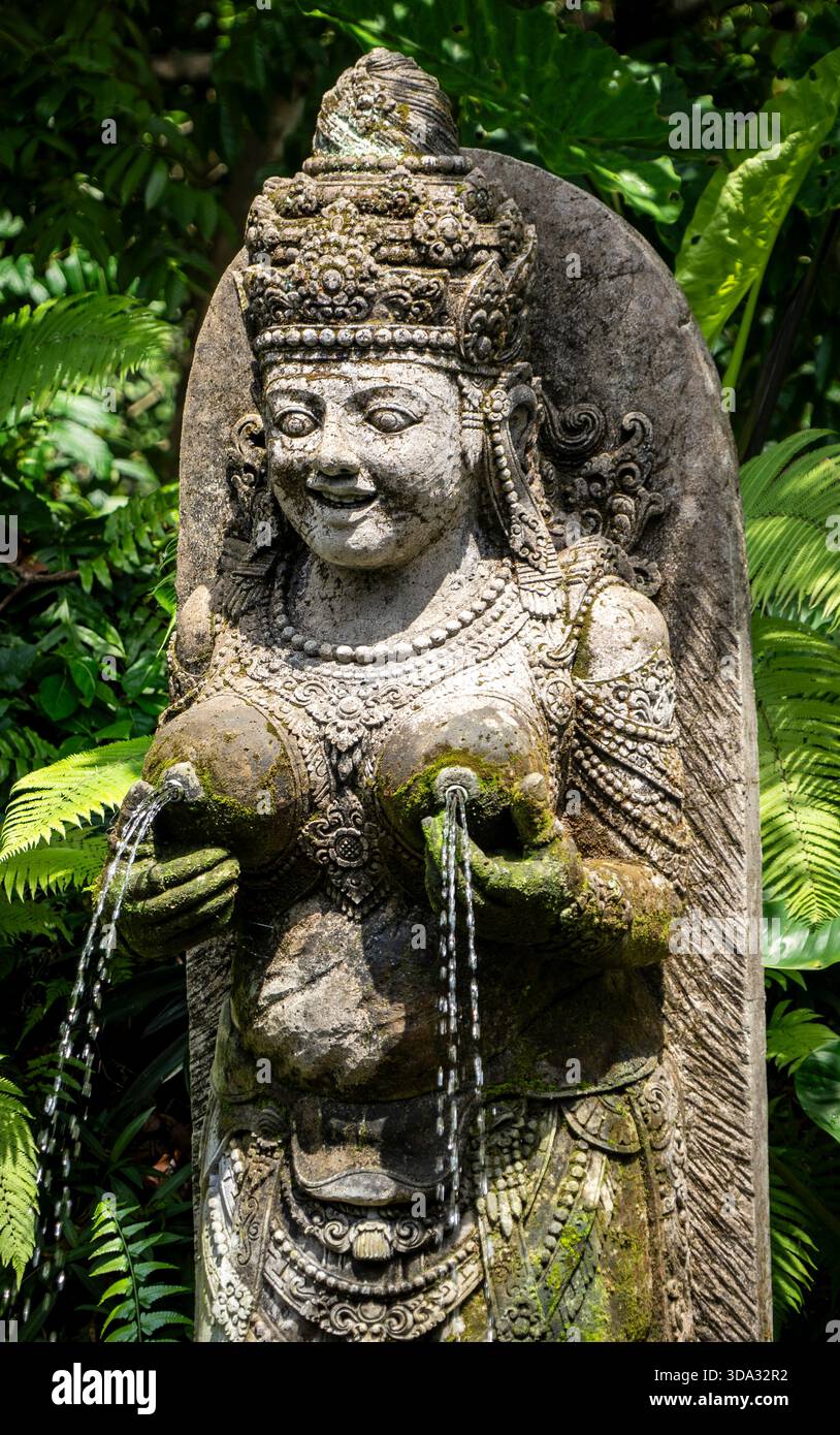 Antica statua in pietra di una dea con acqua che scorre dalle sue mani, circondata da lussureggianti foglie verdi. Foto Stock