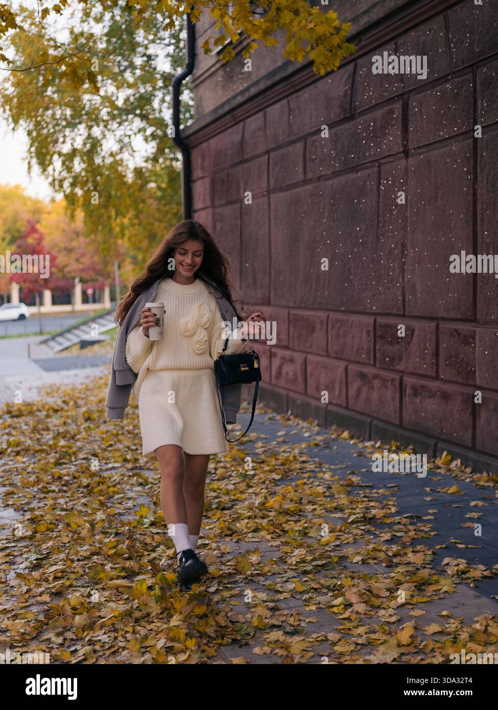 Elegante giovane donna in maglia bianca e giacca grigia che cammina allegramente sul marciapiede ricoperta di foglie gialle cadute, tenendo in mano una tazza di caffè e una h nera Foto Stock