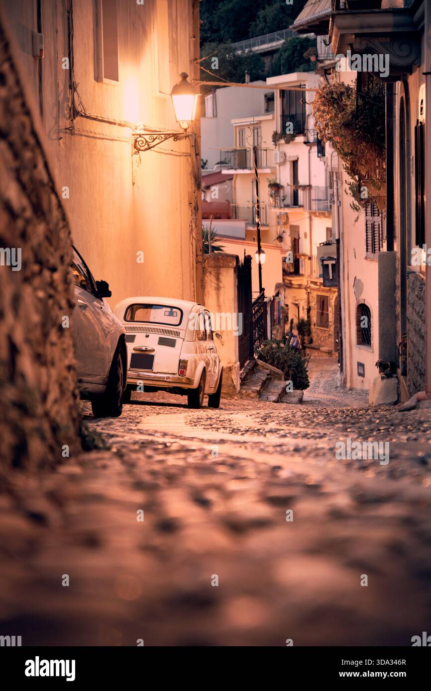 Calda strada di ciottoli serale in una città vecchia mediterranea con una piccola auto d'epoca e luci calde. Foto Stock