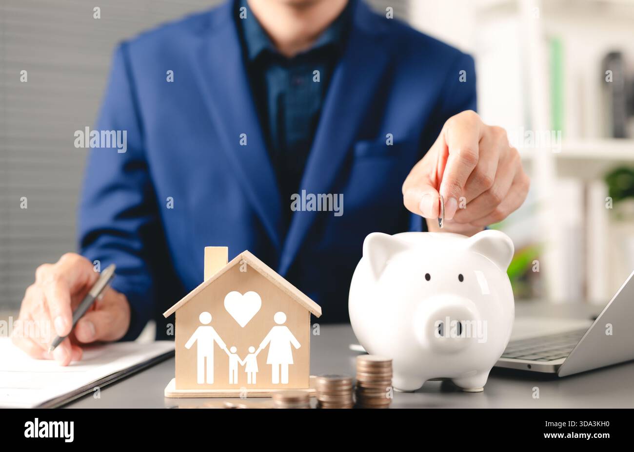Concetto di risparmio economico per la famiglia. L'uomo d'affari deposita monete nel salvadanaio con un modello di casa in legno, simboleggiando la famiglia e l'amore, la pianificazione finanziaria della famiglia Foto Stock