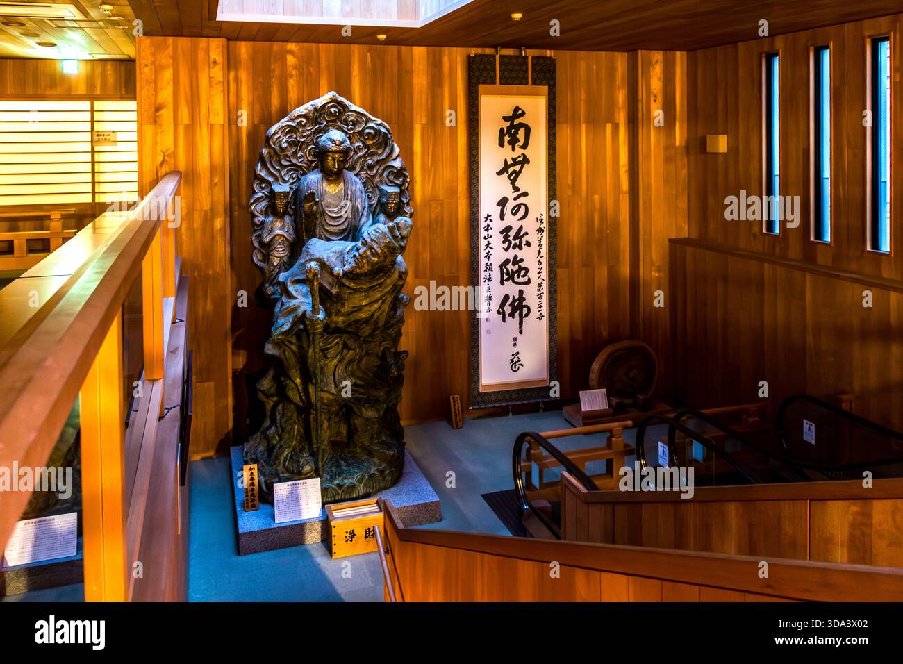 Una grande statua in bronzo di un Buddha seduto (presumibilmente Amida Nyorai) all'interno di un edificio del tempio di Nagano, fiancheggiata da un rotolo calligrafico e da una scala mobile. La giustapposizione della scultura religiosa medievale e della moderna tecnologia costruttiva è caratteristica del tempio Zenkoji (善光寺), uno dei più importanti siti di pellegrinaggio buddista del Giappone, che attrae diversi milioni di visitatori ogni anno. Nagano, Prefettura di Nagano, Giappone Foto Stock