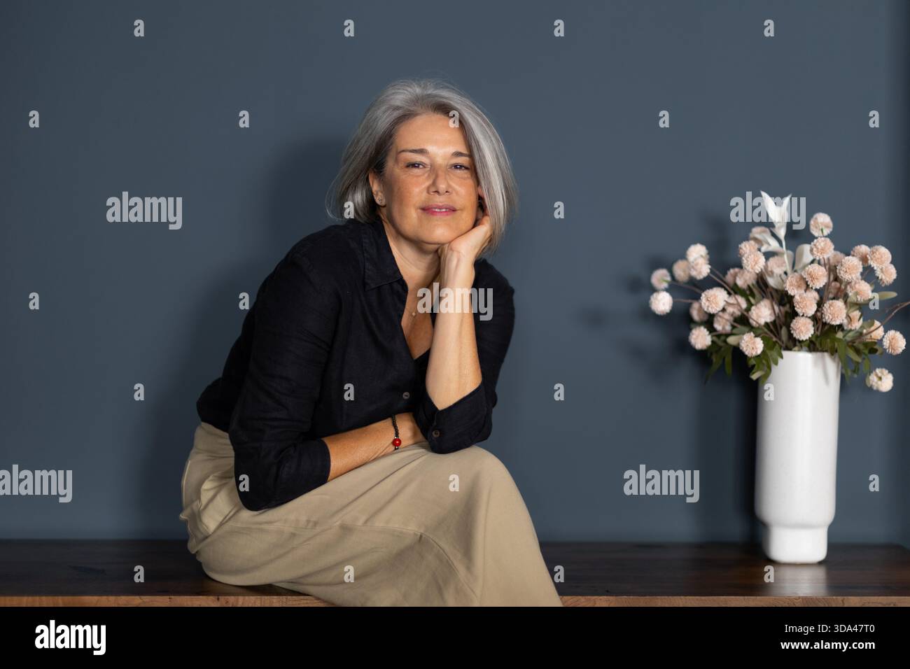 Ritratto di una donna d'affari sorridente e matura seduta su una panchina di legno con la mano sul mento, capelli grigi e fiori in vaso Foto Stock
