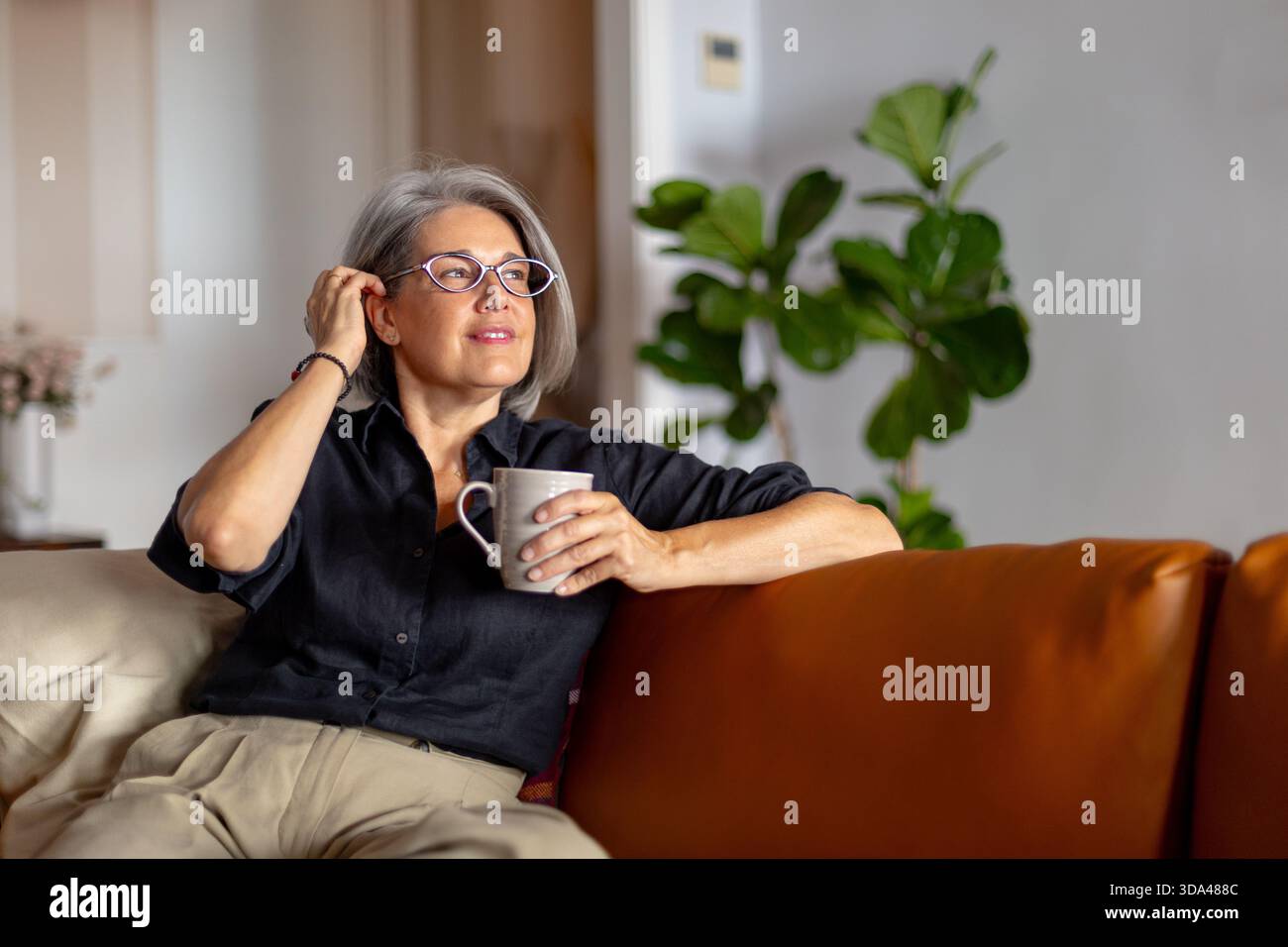 Donna matura dai capelli grigi con occhiali da vista che si gode una tazza di caffè o tè mentre si siede su un comodo divano nel suo moderno soggiorno Foto Stock