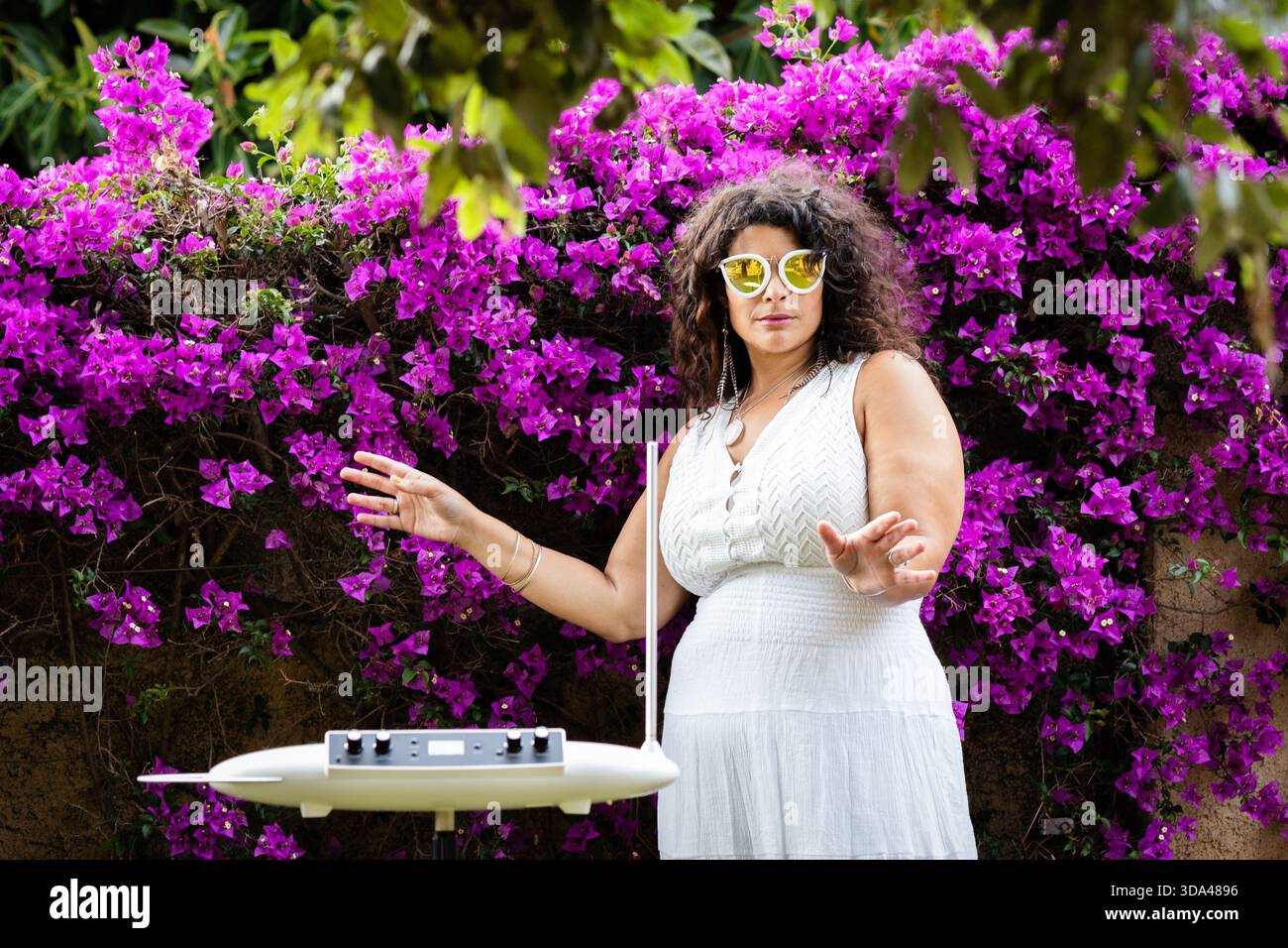 Una donna stupenda in un abito bianco che suona con grazia il theremin in mezzo a splendidi giardini, mescolando eleganza e musica in un ambiente tranquillo Foto Stock