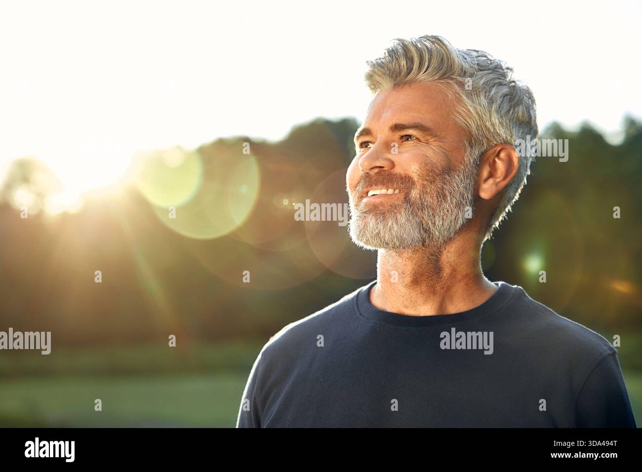 Un uomo sorridente e middleaged, caratterizzato da capelli e barba grigi, si erge all'aperto, illuminato dalla calda luce del tramonto, irradiando positività Foto Stock
