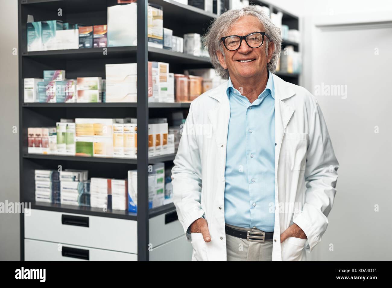 Farmacista anziano sorridente in camice e bicchieri bianchi in piedi in farmacia vicino a scaffali con medicine, sembra sicuro di sé e professionale Foto Stock