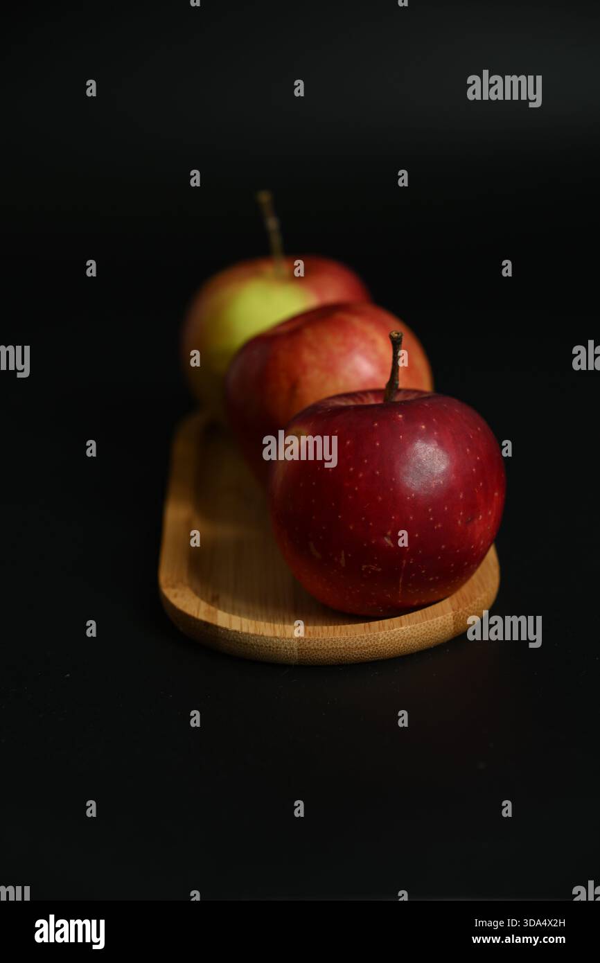 Uno studio STILL Life di tre mele rosse mature disposte verticalmente su un piccolo piatto ovale di legno di bambù su un solido sfondo nero. Selettivo Foto Stock