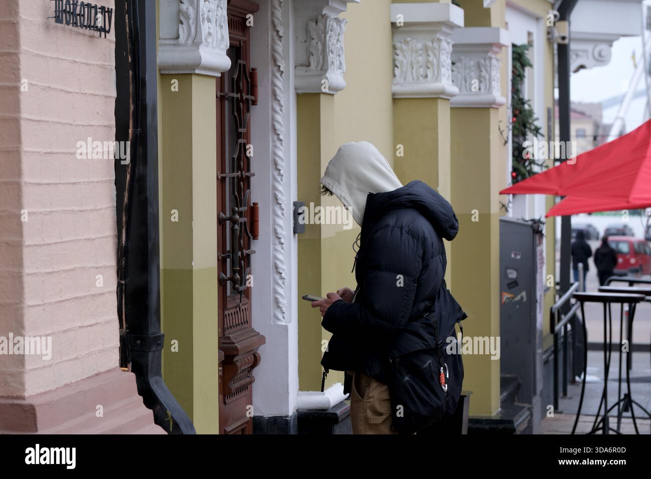 Un giovane con cappuccio si trova accanto a un ingresso elegante, incentrato sul telefono. Podil, Kiev, Ucraina. 8 dicembre 2025. Foto Stock
