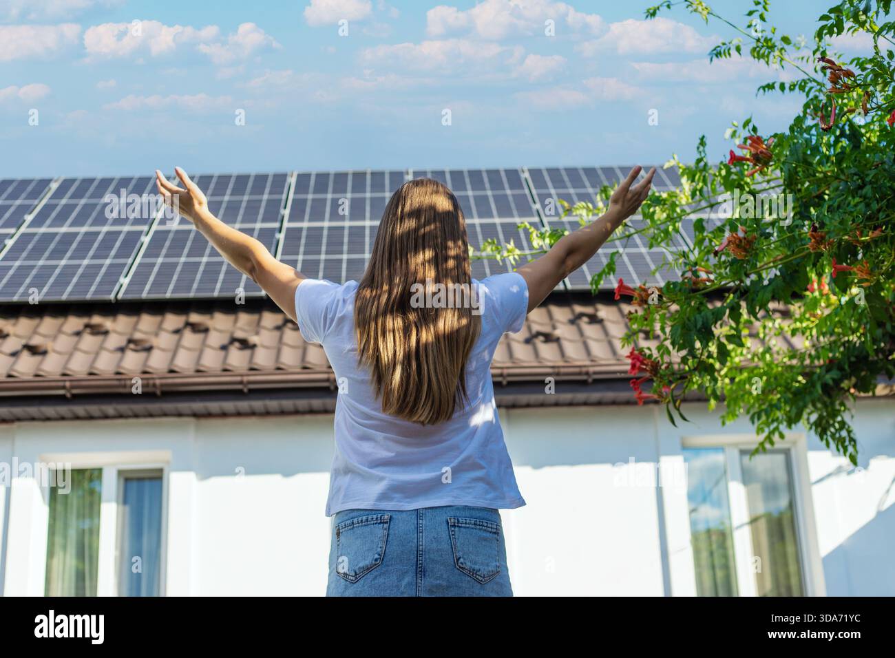 Donna con le braccia alzate di fronte a una casa con pannelli solari e tromba in fiore. Foto Stock